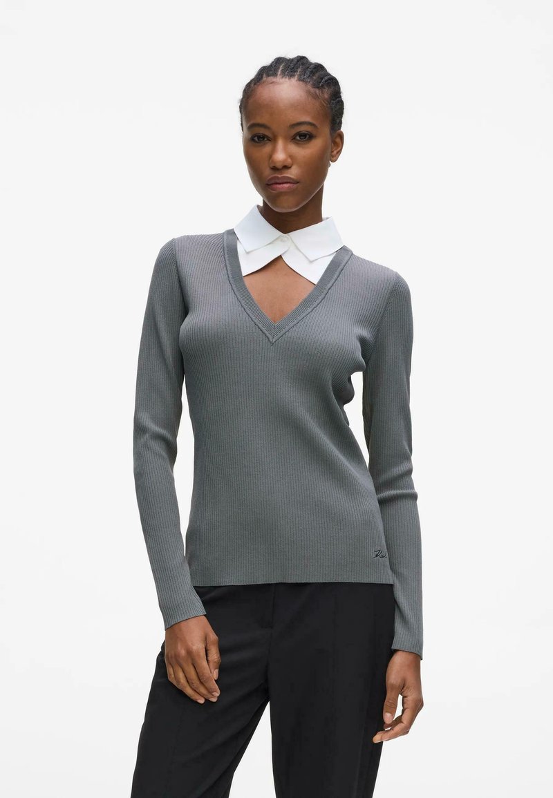 Femme aux cheveux tressés portant un pull gris côtelé à col en V sur un col blanc, associé à un pantalon noir, debout devant un fond blanc uni.