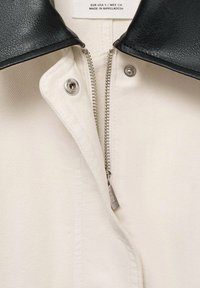 Cream-colored fabric with a black leather collar, featuring a metallic zipper and snap button closure. Textured surface with detailed stitching.