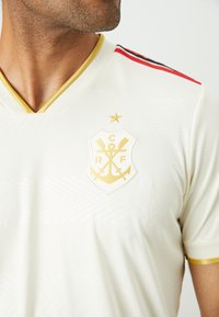 Jersey crema con scollo a V, emblema dorato con ancore incrociate e stelle, strisce rosse e nere sulle spalle e dettagli testurizzati.