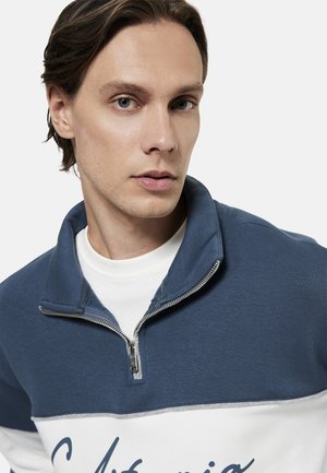 Jonge man met een lichte huid en bruin haar die een blauw-wit quarter-zip sweatshirt draagt met tekst in schrift op de voorkant.