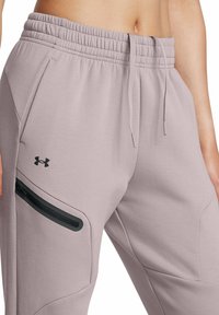 Helle lila Jogginghose mit Kordelzug, gerippten Bündchen und einer seitlichen Reißverschlusstasche mit schwarzen Akzenten. Unter Armour-Logo sichtbar.