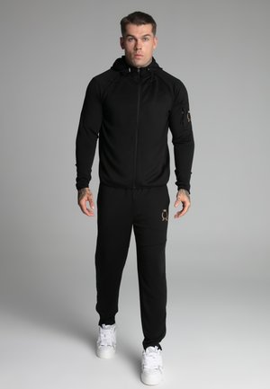 Black zip-up hoodie and pants set with a streamlined design, featuring a logo detail and adjustable drawstrings, paired with white sneakers.