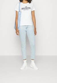 Vit t-shirt med kort ärm och svart "Hollister"-logotyp, ljusblå jeans, vita sneakers och en ljusblå tygkasse.