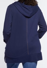 Marinblå luvtröja med rak skärning, med en kontrasterande vertikal sömdetalj mitt på ryggen och sidospår vid fållen.