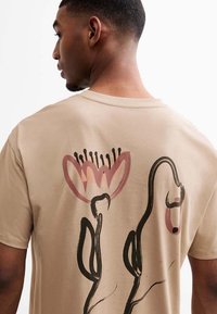 Camiseta de algodón beige con un gran diseño floral abstracto en rosa y negro en la parte posterior, con líneas y formas distintivas.