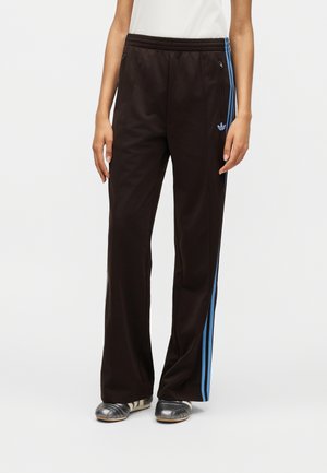 Pantaloni della tuta neri con strisce laterali blu e logo Adidas, indossati con scarpe slip-on argento e top bianco, mostrati dalla vita ai piedi.