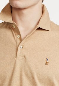 Polo de color marrón claro con una textura suave, que presenta una tapeta de tres botones y un logotipo de un jugador de polo montado en un caballo, bordado en color.