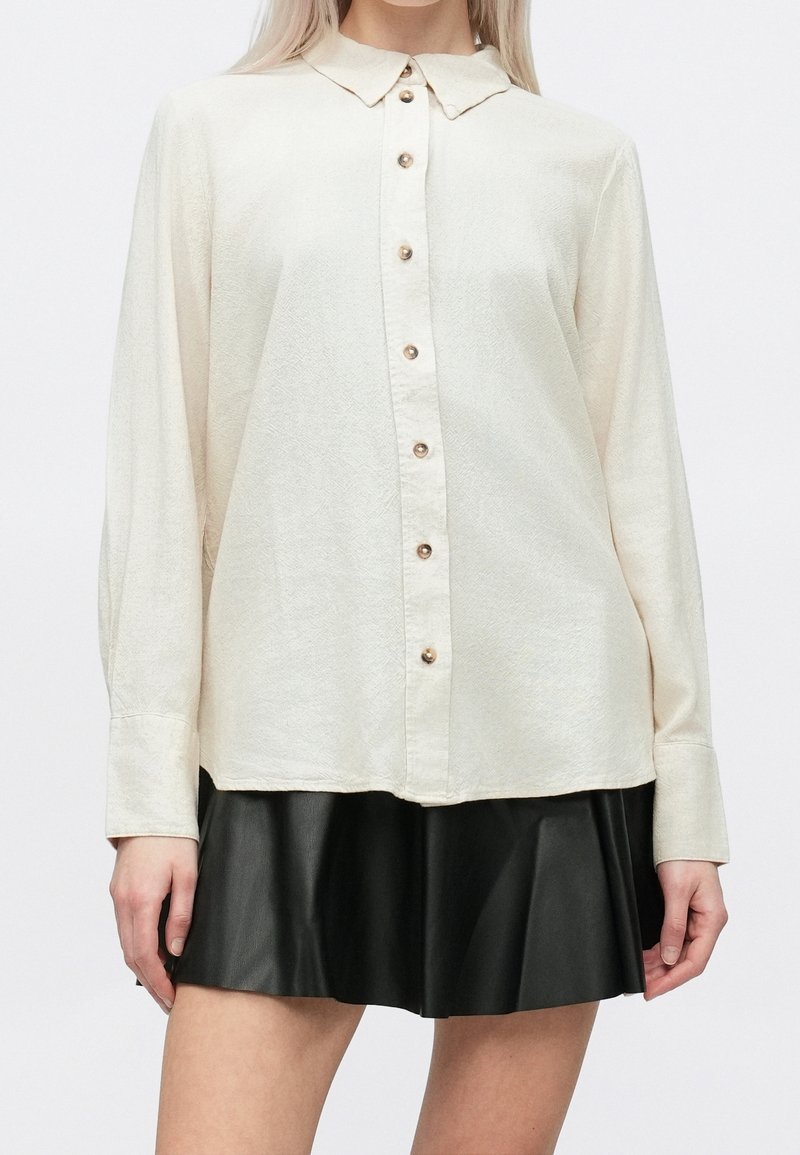 Femme portant une chemise crème à manches longues boutonnée, associée à une mini-jupe plissée noire, debout devant un fond uni.
