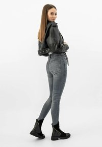 Černá syntetická kožená bombera přes bílý top, doplněná šedými skinny džíny s vysokým pasem a černými kotníkovými botami se zipem.