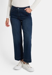 Femme portant un jean droit bleu marine et des baskets blanches, avec une main dans la poche et un haut à manches longues blanc.