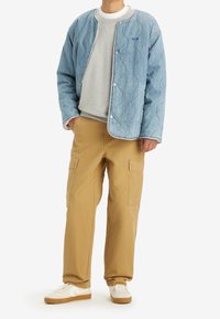 Niebieska pikowana kurtka dżinsowa z zatrzaskami, szary sweter pod spodem, beżowe spodnie cargo z kieszeniami i białe trampki z gumową podeszwą.