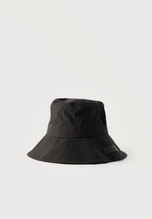 Zwarte stoffen bucket hat met brede, gestikte rand en klein rechthoekig label aan de zijkant, weergegeven op een effen witte achtergrond.