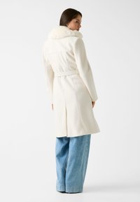 Langer weißer Mantel mit strukturiertem Finish und einem plüschigen Fellkragen. Der Mantel hat eine taillierte Taille und eine gerade Silhouette, kombiniert mit hellblauen weit geschnittenen Jeans.