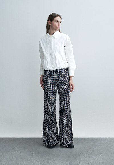 Femme portant un chemisier blanc à manches longues avec un col large et un pantalon large bleu marine à motifs, debout sur un sol gris devant un mur gris clair.