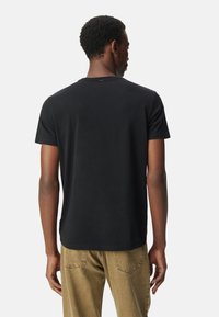 Uomo visto da dietro che indossa una semplice maglietta nera a maniche corte e jeans marrone chiaro, in piedi contro uno sfondo bianco.