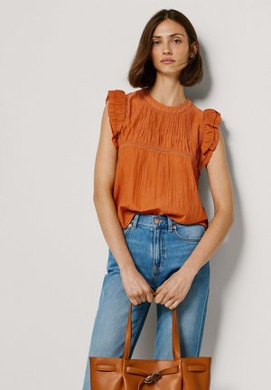 Kvinde med kort brunt hår iført en orange ærmeløs bluse med flæser og lyseblå jeans, der holder en brun lædertaske foran sig.