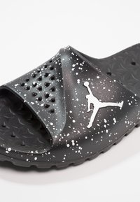 Musta liukusandaali, jossa on valkoinen maaliroiskepattern ja reikänauha, jossa on valkoinen siluetti koripallopelaajasta.