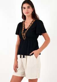Zwarte blouse met korte pofmouwen, met een oranje geborduurde detailing langs de halslijn en een aansluitende taille, gecombineerd met beige shorts.