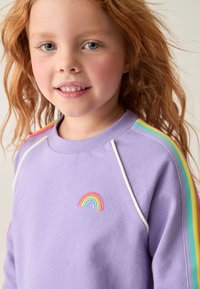 Lila Sweatshirt mit weißen Akzenten und Regenbogen-Stickerei auf der Brust. Verfügt über mehrfarbige gestreifte Details an den Ärmeln.