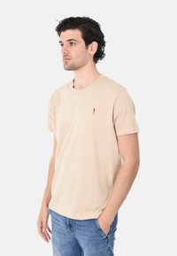 T-shirt in cotone beige con maniche corte, colletto a girocollo e un piccolo ricamo di un peperoncino rosso sul lato sinistro del petto. Indossato con jeans blu.