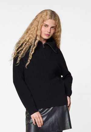 Pull en tricot noir à côtes, avec un col haut et une demi-fermeture éclair. Les manches sont longues et la coupe est décontractée. Associé à une jupe en cuir.