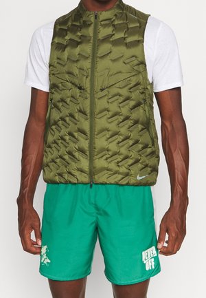 Mand iført olivengrøn polstret vest over hvid kortærmet skjorte og lysegrønne shorts med teksten "NEVER OFF" på venstre ben.