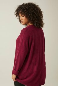 Maglione oversize bordeaux con una texture morbida, dotato di borchie decorative sulle spalle, fessure laterali e polsini arrotolati.