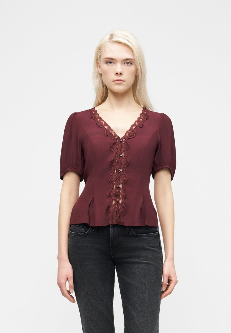 Γυναίκα με ίσια ξανθά μαλλιά φοράει μπορντό μπλούζα με δαντέλα και κουμπιά, συνδυασμένη με ξεθωριασμένο μαύρο τζιν, στέκεται μπροστά σε λευκό φόντο.