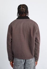 Brun bomuldsjakke med en kordfløjlskrave, oversized design og trykknapdetaljer, set fra ryggen. Tekstureret stof og afslappet pasform.