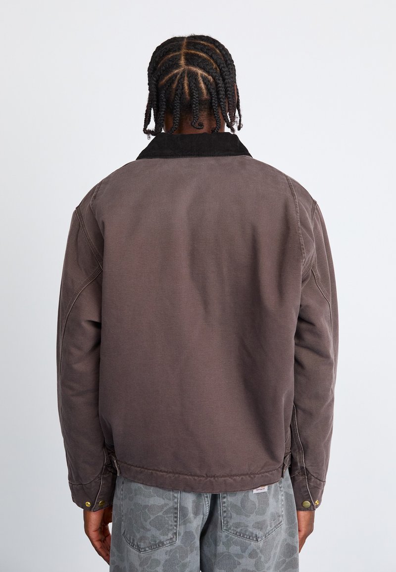 Bruine katoenen jas met een corduroy kraag, oversized ontwerp en drukknoopaccenten, gezien vanaf de achterkant. Gelaagd materiaal en een relaxte pasvorm.