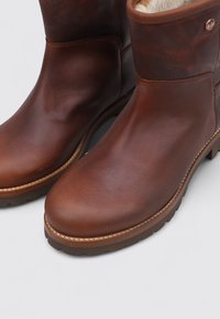 Botas de cuero marrón hasta el tobillo con un acabado suave, puntera redonda y forro acolchado. Cuenta con una suela de goma y detalles cosidos a lo largo del borde.