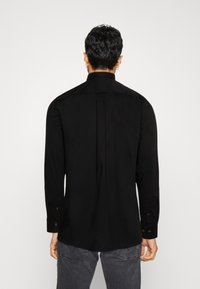 Homme aux cheveux courts et foncés portant une chemise noire à manches longues et un jean gris foncé, vu de dos sur un fond blanc uni.