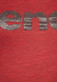 Rød stof med en tekstureret overflade, der har store, glitrende sølvbogstaver, som staver "ence" på forsiden. Glat, afslappet design.