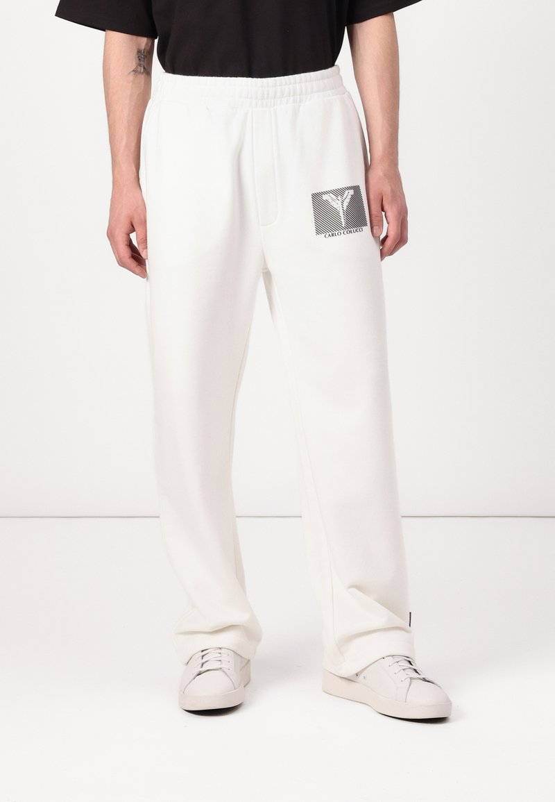 Witte trainingsbroek met een elastische tailleband, zijzakken en een grafisch logopatch in zwart en grijs op de linker dij. Soepele textuur.