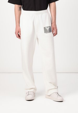 White sweatpants with an elastic waistband, side pockets, and a graphic logo patch in black and gray on the left thigh. Smooth texture.