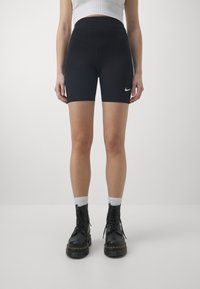 Donna che indossa pantaloni corti neri Nike, calze bianche e stivali neri con lacci, in piedi contro uno sfondo chiaro e semplice.
