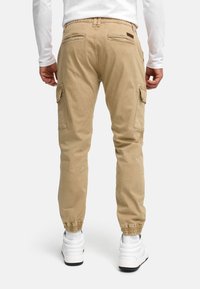 Homme portant un pantalon cargo beige et des baskets blanches, debout de dos avec les mains détendues le long du corps, sur un fond blanc.
