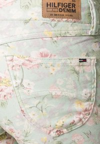 Jeans de denim con estampado floral en verde menta, que presentan flores rosas y amarillas, detalle en el bolsillo trasero y una etiqueta de marca de cuero marrón.
