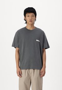 Camiseta de algodón gris con cuello redondo, mangas cortas y un pequeño logo blanco en el lado izquierdo del pecho. El corte es relajado y casual.