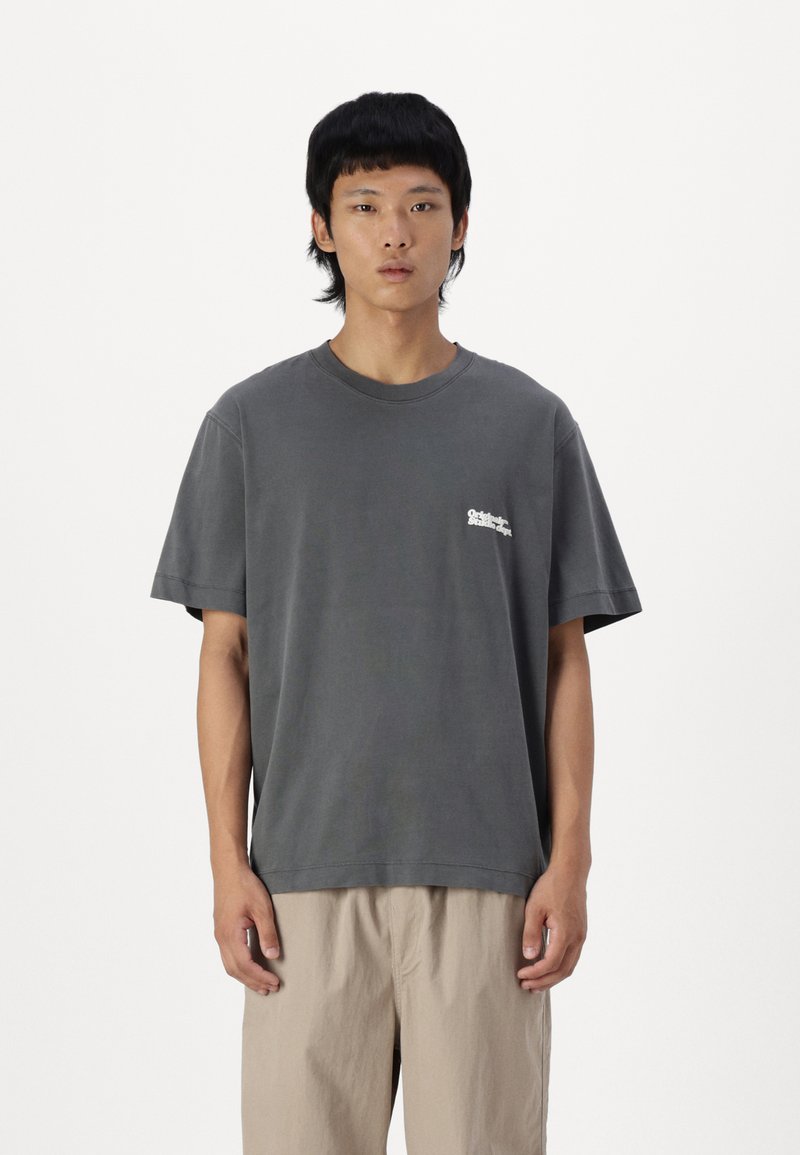 Camiseta de algodón gris con cuello redondo, mangas cortas y un pequeño logo blanco en el lado izquierdo del pecho. El corte es relajado y casual.
