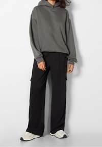 Persona che indossa un ampio hoodie grigio, pantaloni neri a gamba larga con tasche laterali e sneakers bianche, in piedi contro uno sfondo bianco neutro.