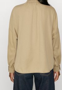 Blouse beige à manches longues avec un empiècement froncé au dos, coupe ample et poignets boutonnés, associée à un jean en denim bleu foncé.