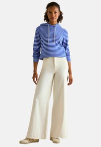 Žena stojí oblečená v modro-bielom pruhovanom mikinovom svetri, širokých krémových nohaviciach a béžových teniskách na jednoduchom svetlom pozadí.