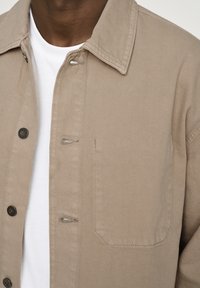 Beige, strukturiertes Jacket mit umklappbarem Kragen, Knopfschlägen und einer Brusttasche. Getragen über einem weißen Hemd, das das geschichtete Design zur Geltung bringt.