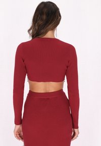 Vinröd ribbad croptop med långa ärmar och öppen rygg, med en texturerad stickad design. Justerad midja kompletterar stilen.