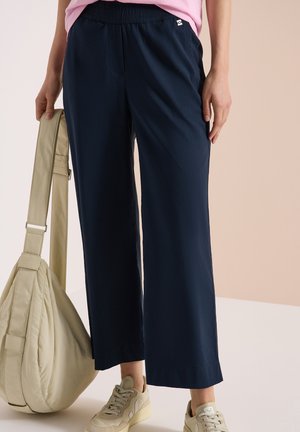 Personne portant un pantalon large bleu marine et des baskets beiges, tenant un grand sac à épaule beige avec une sangle réglable.