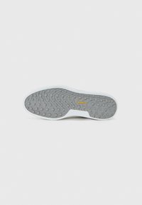 Semelle en caoutchouc, présentant un motif texturé gris avec des accents de grip circulaires, bordée d'un bord blanc ; logo de la marque en jaune.