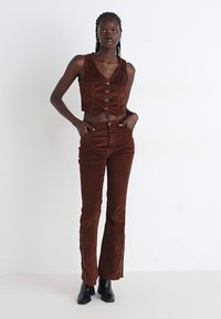 Brown corduroy cropped vest with buttons paired with matching flared pants, featuring a smooth texture and fitted design, worn with black boots.