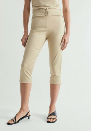 Mujer con pantalones beige recortados de cintura alta y aberturas laterales, combinados con sandalias negras de tacón bajo, de pie frente a un fondo liso.