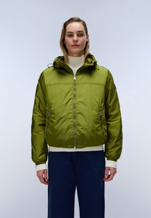 Femme porte une veste zippée verte sur un col roulé blanc et un pantalon bleu foncé, debout devant un fond gris clair.